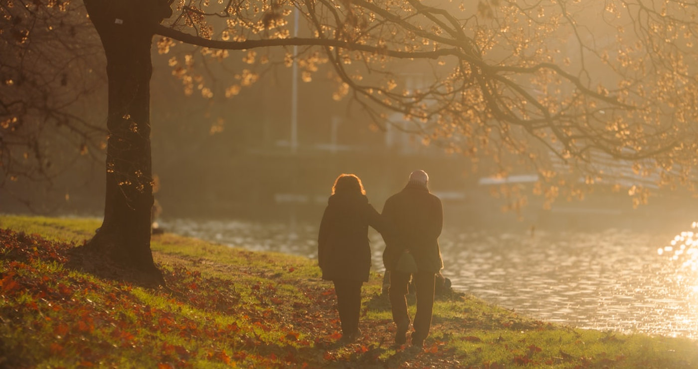 An older couple walking together beside a river under an autumn tree, backlit by low sun.